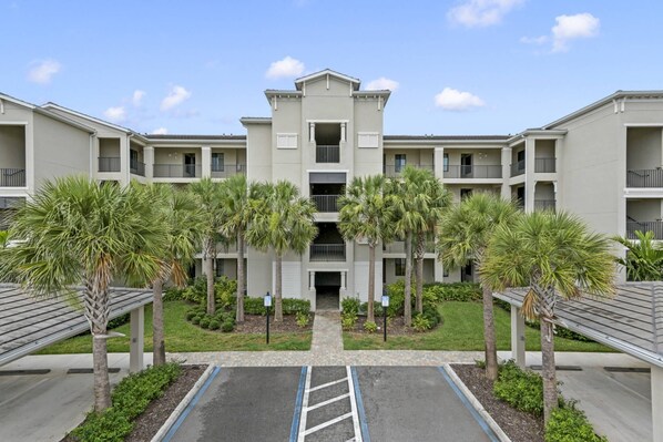 A multi-story residential building with a central entrance, surrounded by palm trees and a parking area.