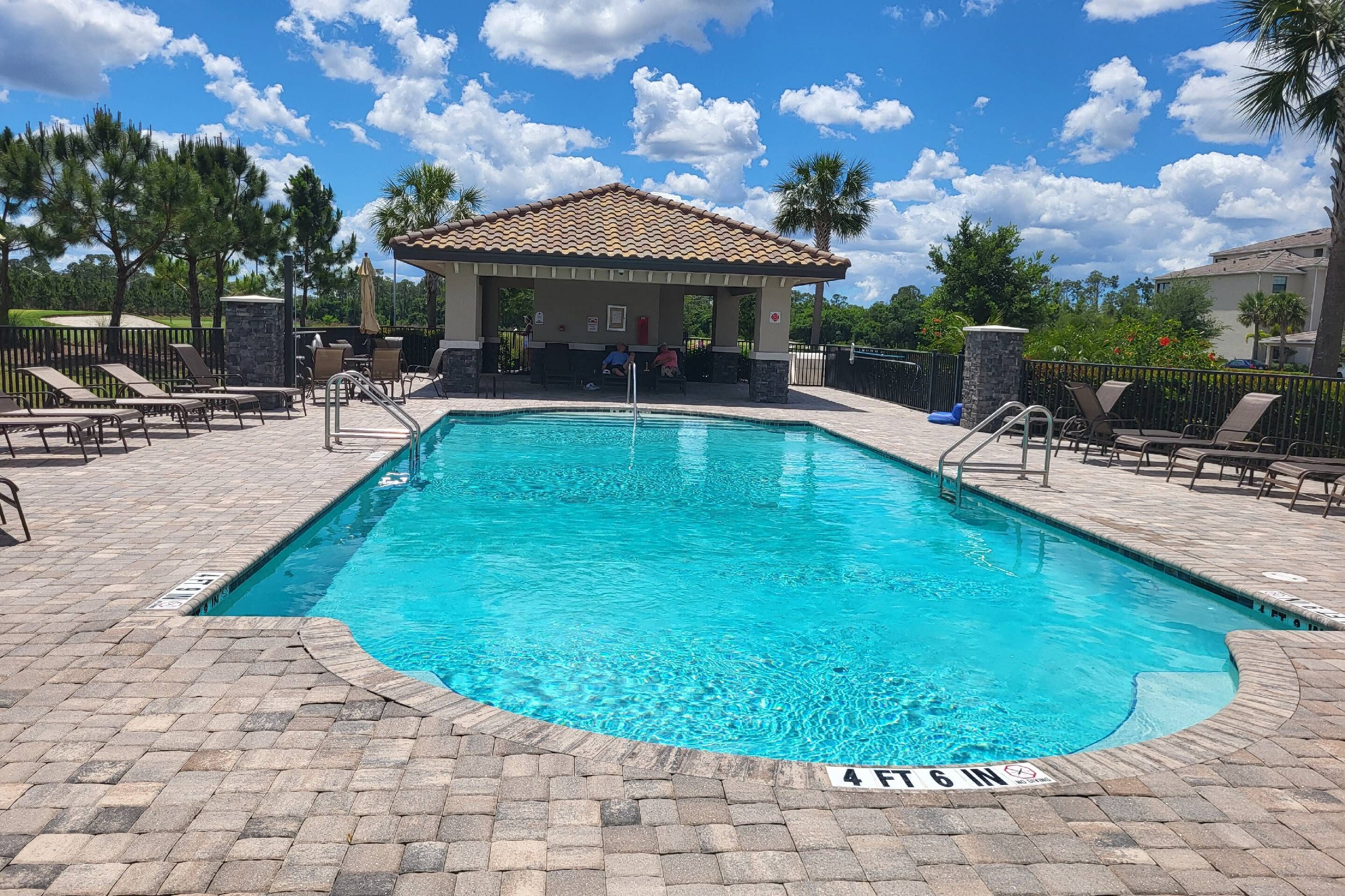A swimming pool with a tiled deck, a covered seating area, and a clear sky.