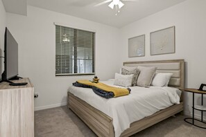 A bedroom with a bed, a television, a window with blinds, and two wall-mounted artworks.