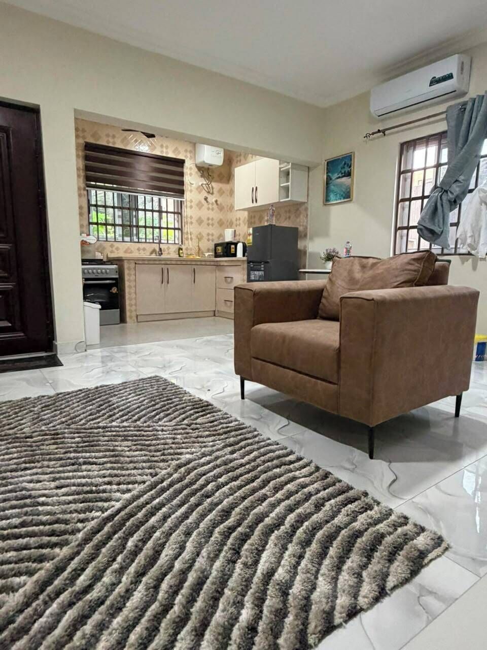 Uma sala de estar com sofá, tapete e cozinha ao fundo, além de um aparelho de ar condicionado.