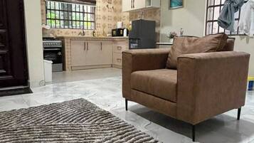 Uma sala de estar com sofá, tapete e cozinha ao fundo, além de um aparelho de ar condicionado.