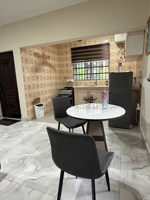 Un comedor con una mesa redonda y cuatro sillas, una pequeña cocina con estufa y lavabo, y una ventana con barrotes.