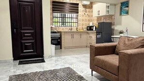 Una sala de estar con sofá y alfombra estampada, una puerta de madera oscura, una cocina con armarios y electrodomésticos, y un ventilador de techo.