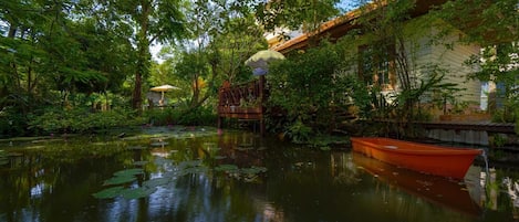 A serene pond with a wooden boat, surrounded by lush greenery and a traditional bridge.
