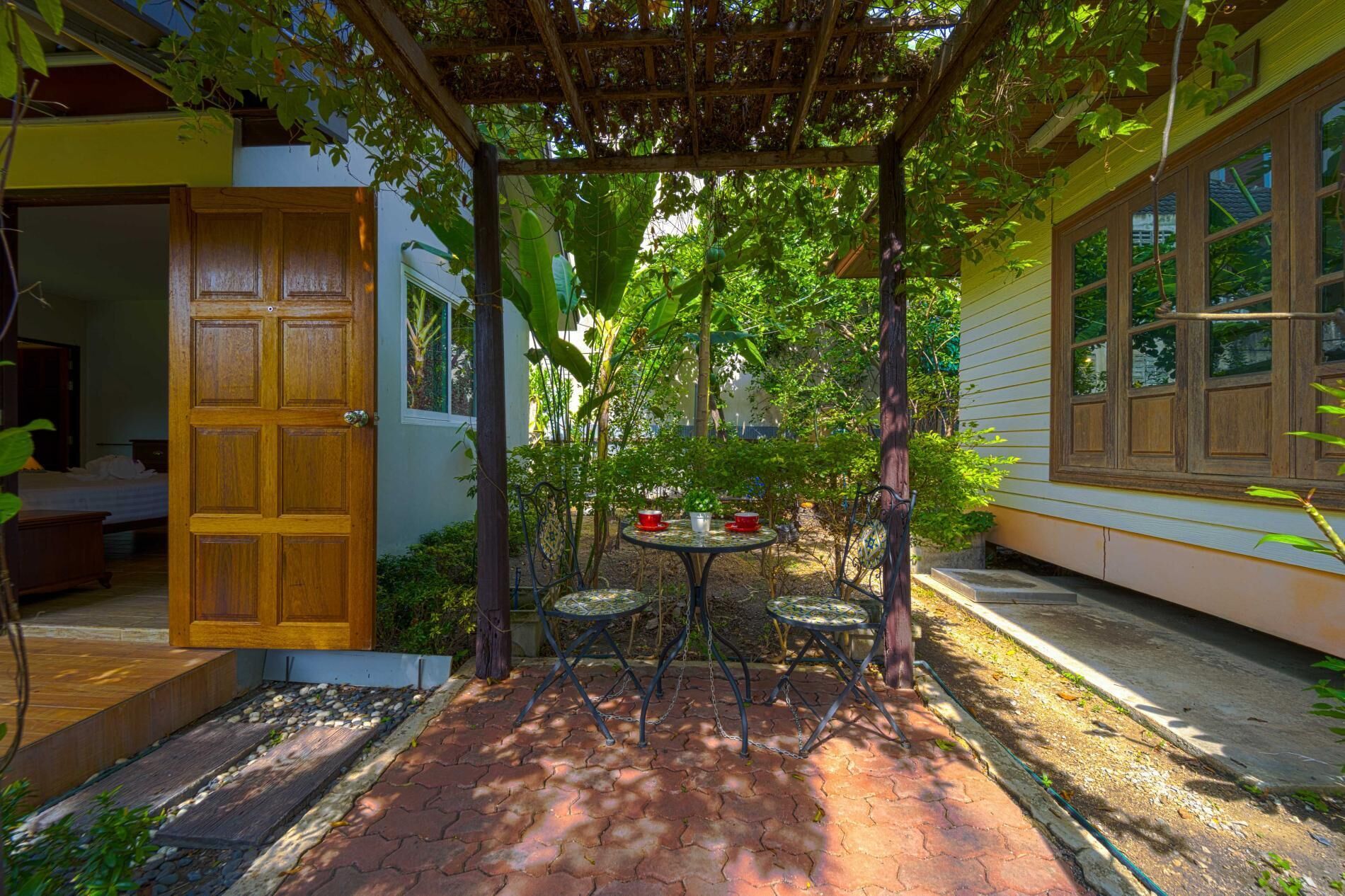 Un porche cubierto con una puerta de madera, una mesita con sillas y un jardín con plantas, incluyendo una maceta.