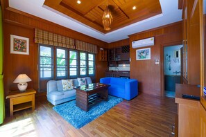 A cozy living room with a wooden ceiling, a blue sofa, a coffee table, and a kitchen area with cabinets and a sink. - baansuanmuenbuppha (Hua Hin)