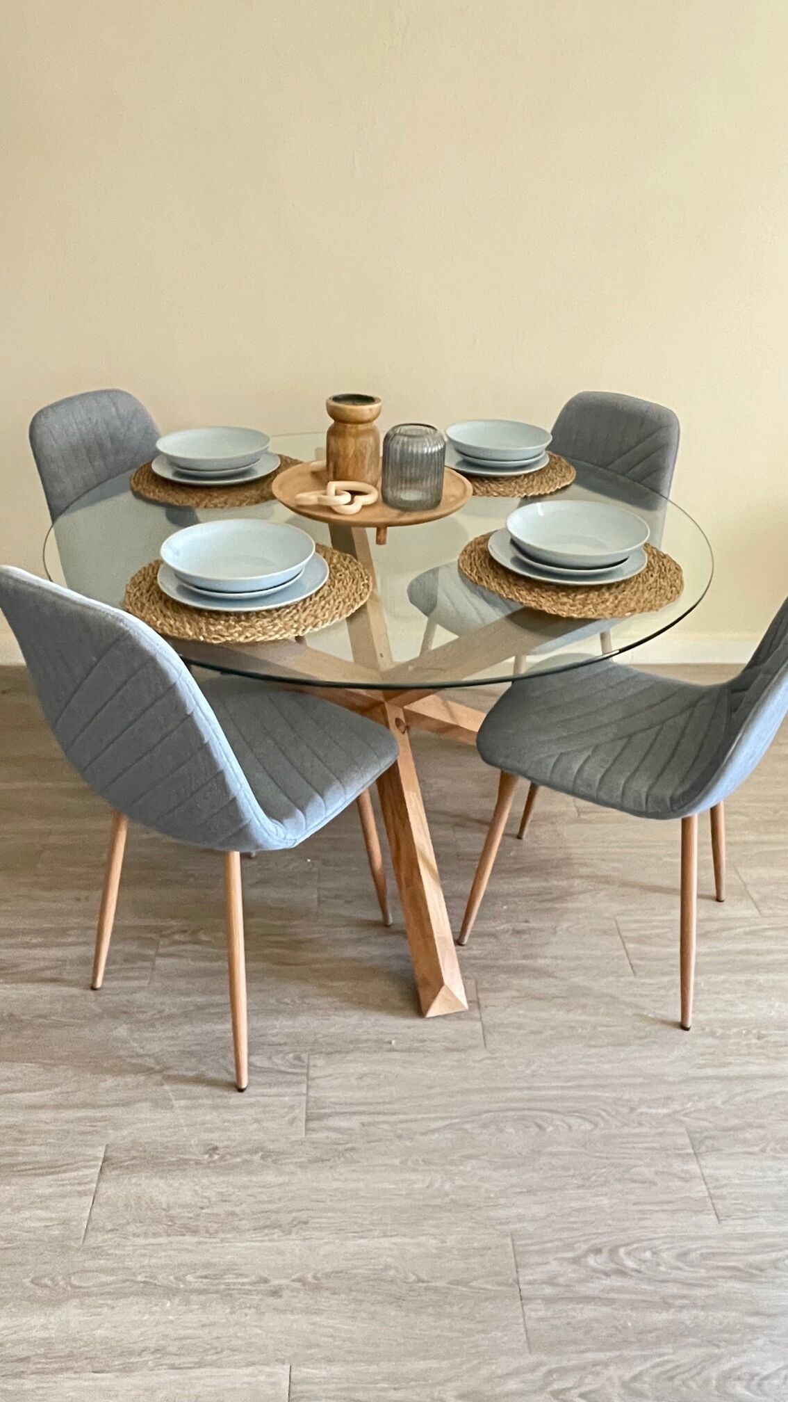 A round glass table with woven placemats, grey upholstered chairs, and stacked plates.