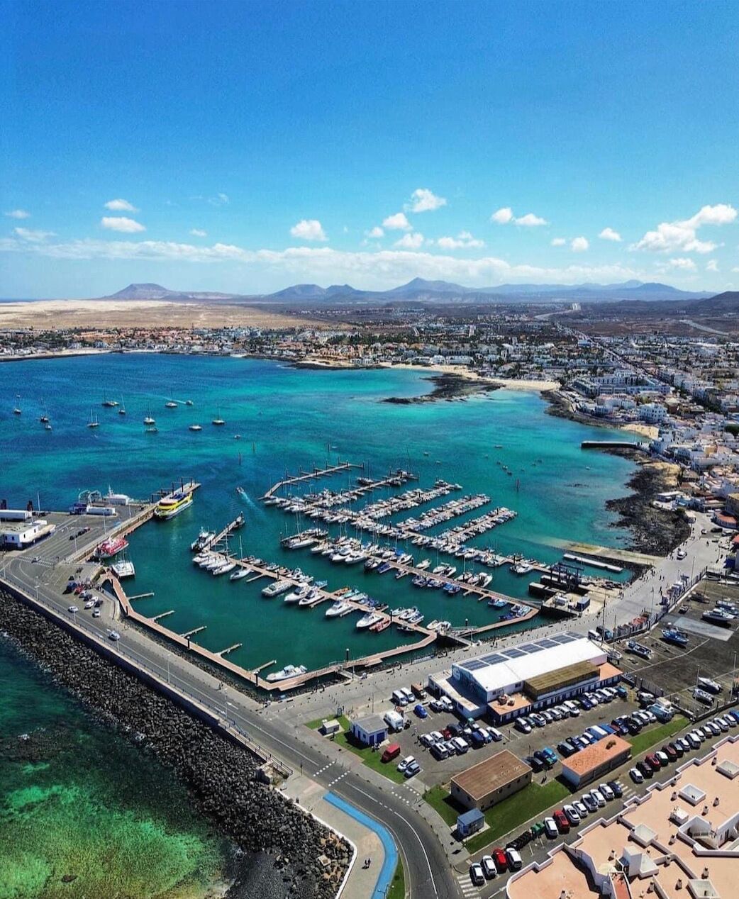 An aerial view of a coastal marina with boats, a road, and buildings.