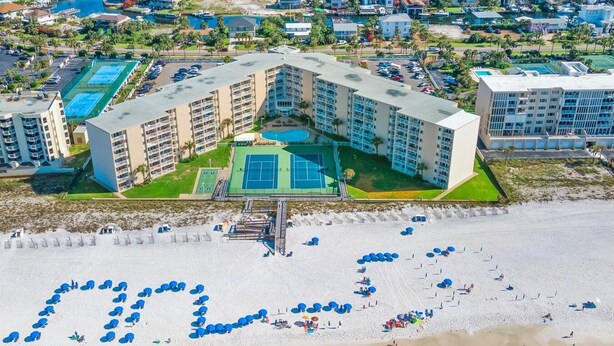 Un hotel frente a la playa con varios pisos, una cancha de tenis y una playa con sombrillas dispuestas en forma de corazón.