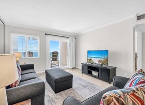 Ein modernes Wohnzimmer mit einem Flachbildfernseher, grauen Sofas, einem Couchtisch und Blick ins Freie durch große Fenster.