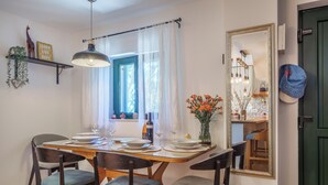 Comedor con mesa de madera dispuesta para cuatro, lámpara colgante, ventana con cortinas y un espejo en la pared.