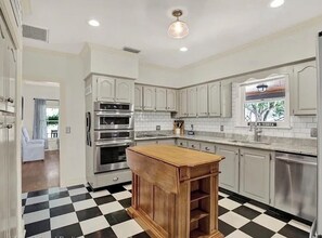 Una cocina con armarios blancos, electrodomésticos de acero inoxidable, una isla de madera y piso a cuadros blanco y negro.