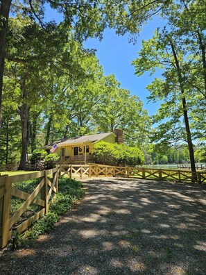 A yellow cabin with a porch, surrounded by trees and a gravel path. - Waterfront Retreat with Private Dock (Heathsville)