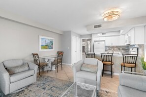 A modern living room with grey sofas, a glass coffee table, and a dining area with chairs and a table. - 2BR Beachfront corner condo w private beach & hot tub ~ Sleeps 8 (Gulf Shores)