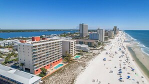 A beachfront hotel with multiple floors, a swimming pool, and a beach with umbrellas and people. - 2BR Beachfront corner condo w private beach & hot tub ~ Sleeps 8 (Gulf Shores)