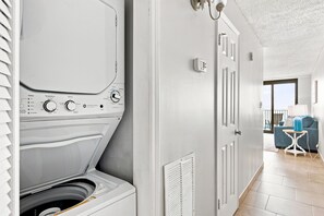 Uma lavanderia com máquina de lavar e secadora, uma porta branca e vista para uma sala de estar com sofá azul e luminária.