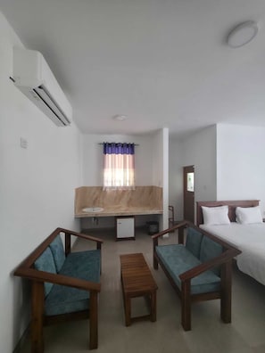Una habitación de hotel con cama, dos sillones, una mesita, ventana con cortinas, lavamanos y aire acondicionado.