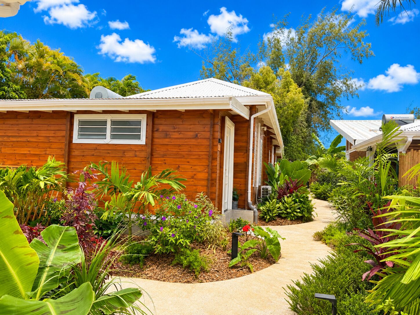 Une maison en bois avec une fenêtre blanche, entourée d’une végétation tropicale luxuriante et sous un ciel bleu azur.
