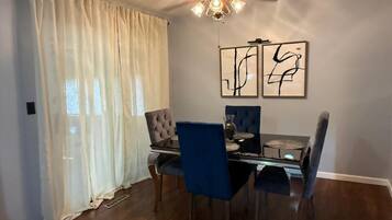 A dining area with a glass table, blue upholstered chairs, a ceiling fan, and abstract wall art.