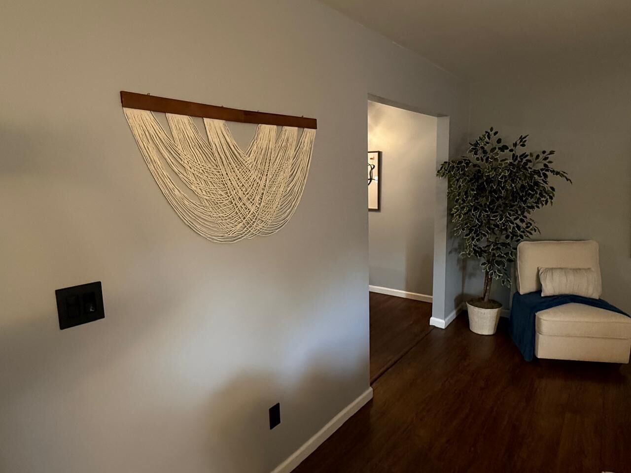 Un pasillo decorado con un macramé colgado en la pared, una planta en maceta y una silla.