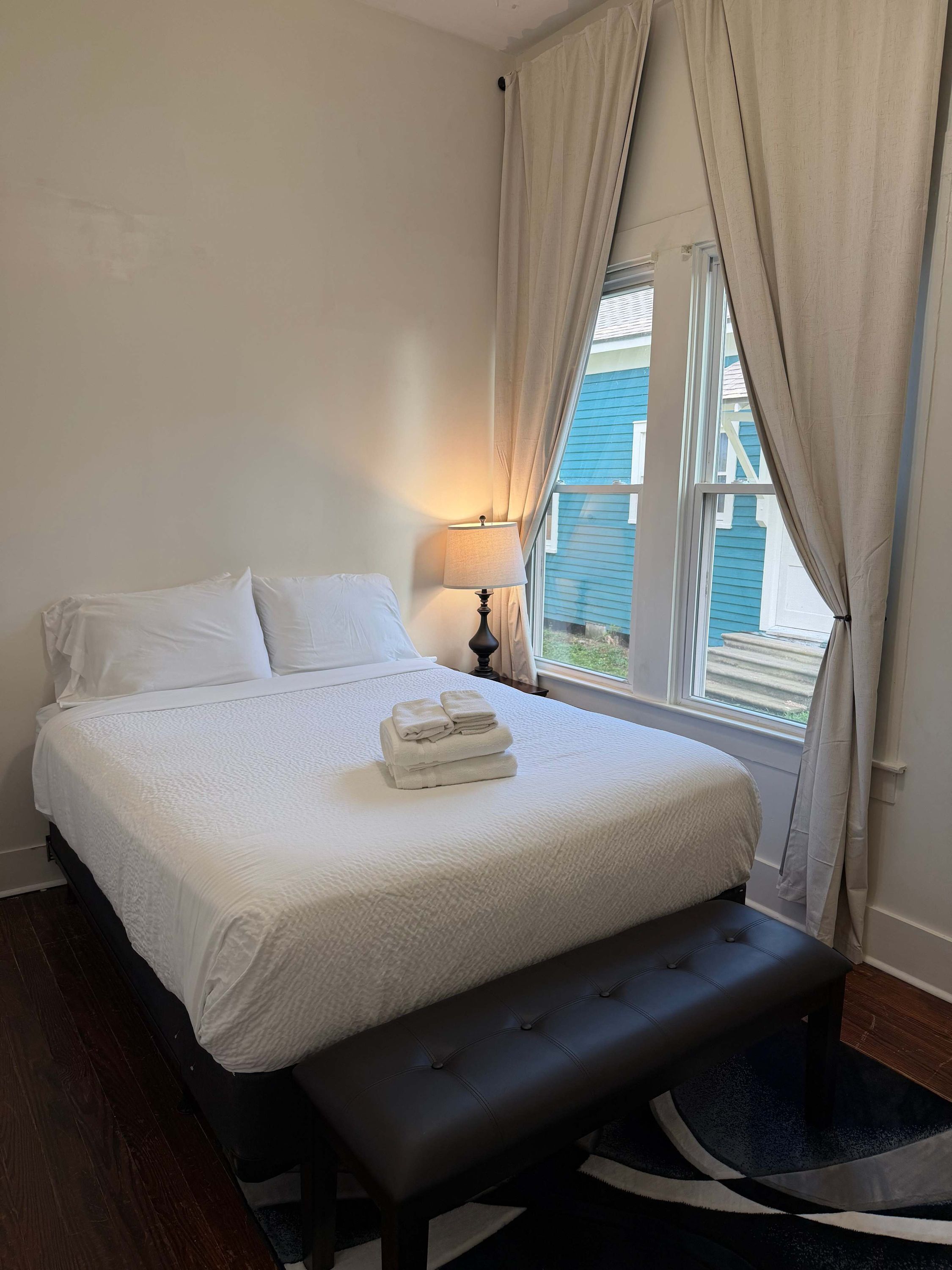 A neatly made bed with white linens and a folded towel, a dark leather bench, a bedside table with a lamp, and a window with curtains.
