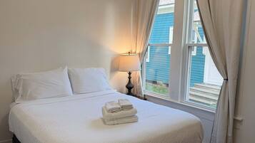 A neatly made bed with white linens and a folded towel, a dark leather bench, a bedside table with a lamp, and a window with curtains.
