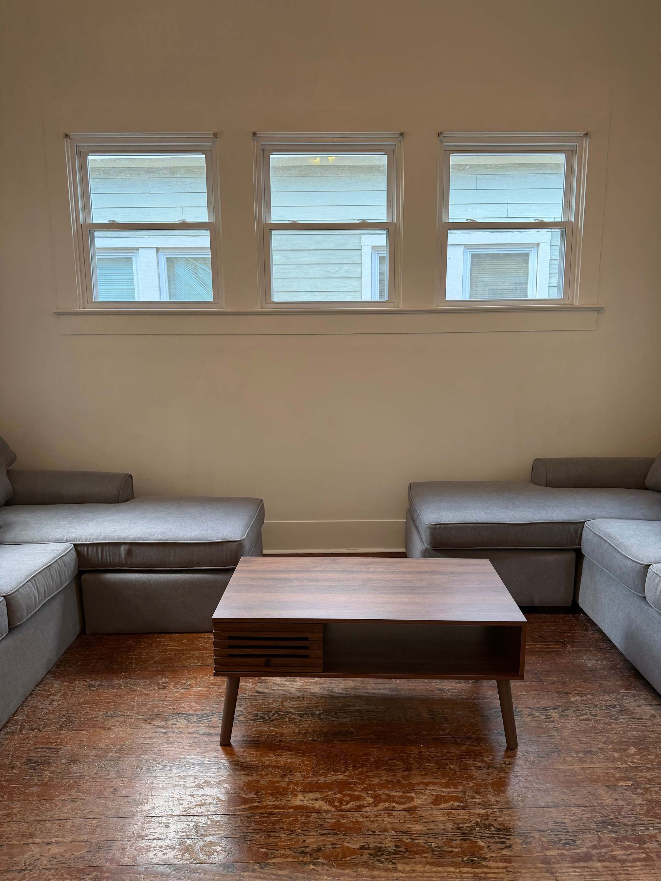 Una sala de estar con un sofá modular, una mesa de centro de madera y tres ventanas con persianas.