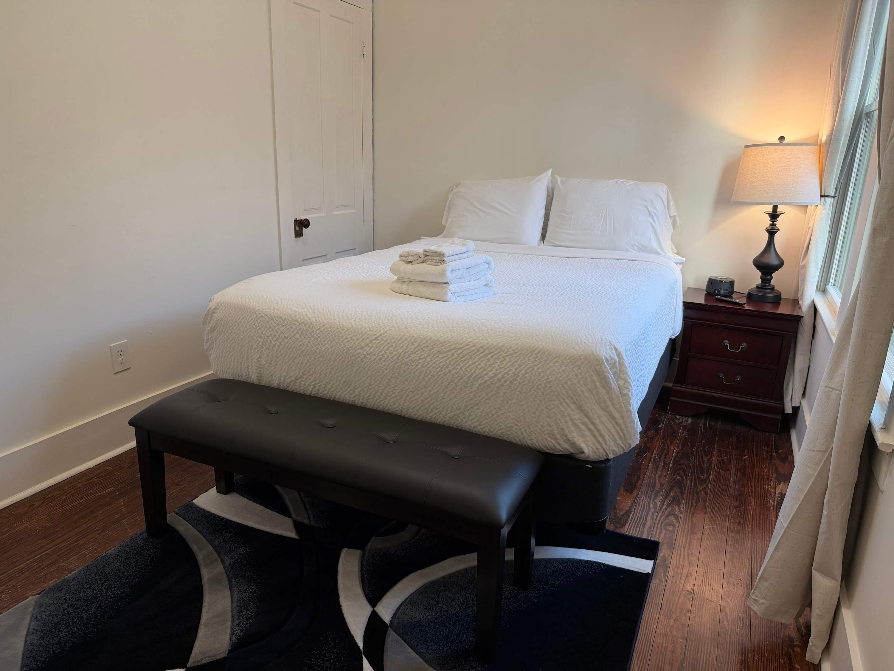 A neatly made bed with a white comforter and pillows, a bedside table with a lamp, and a bench in front of the bed.