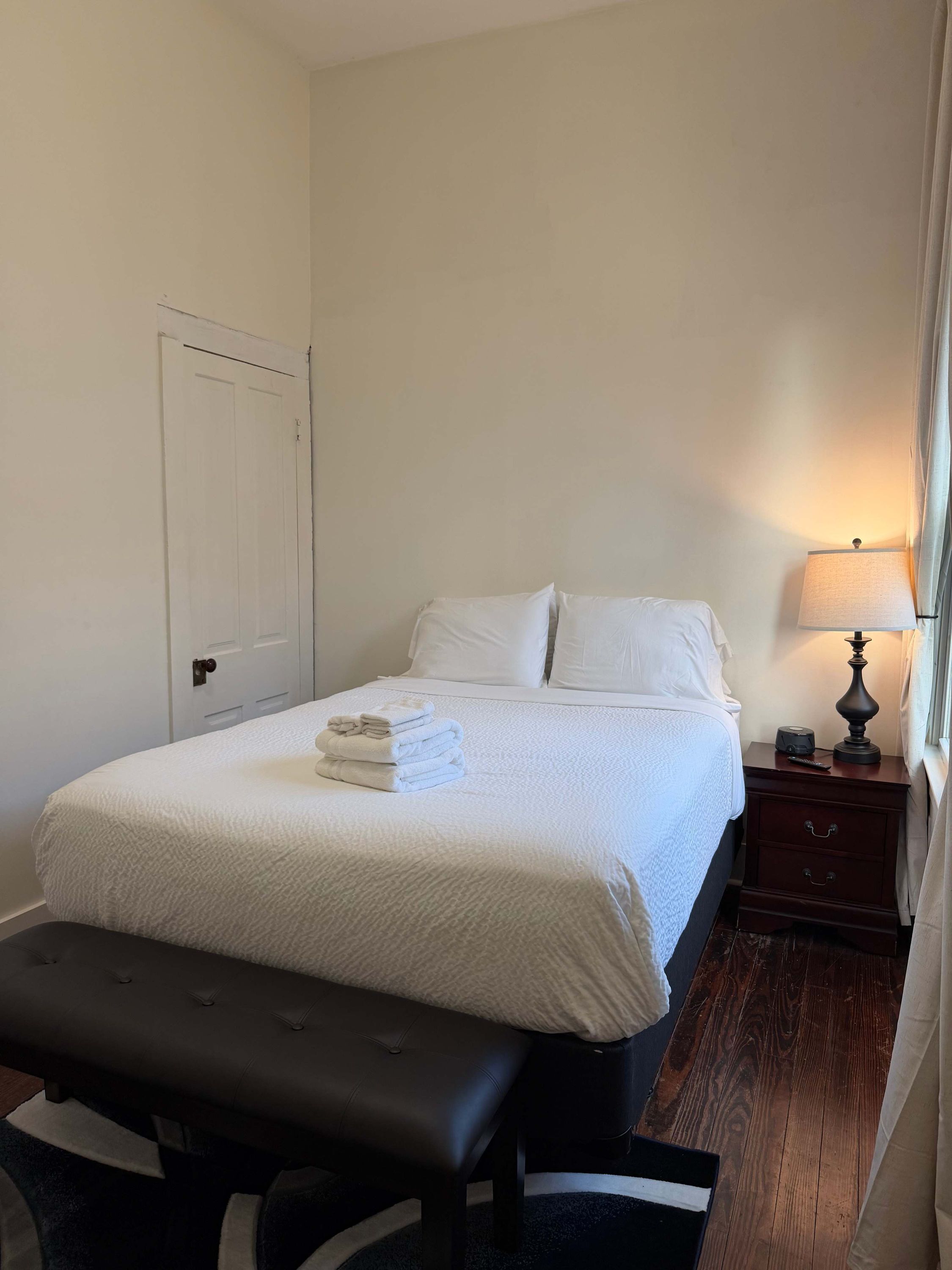 A neatly made bed with white linens, a bedside table with a lamp, and a bench in front of the bed.