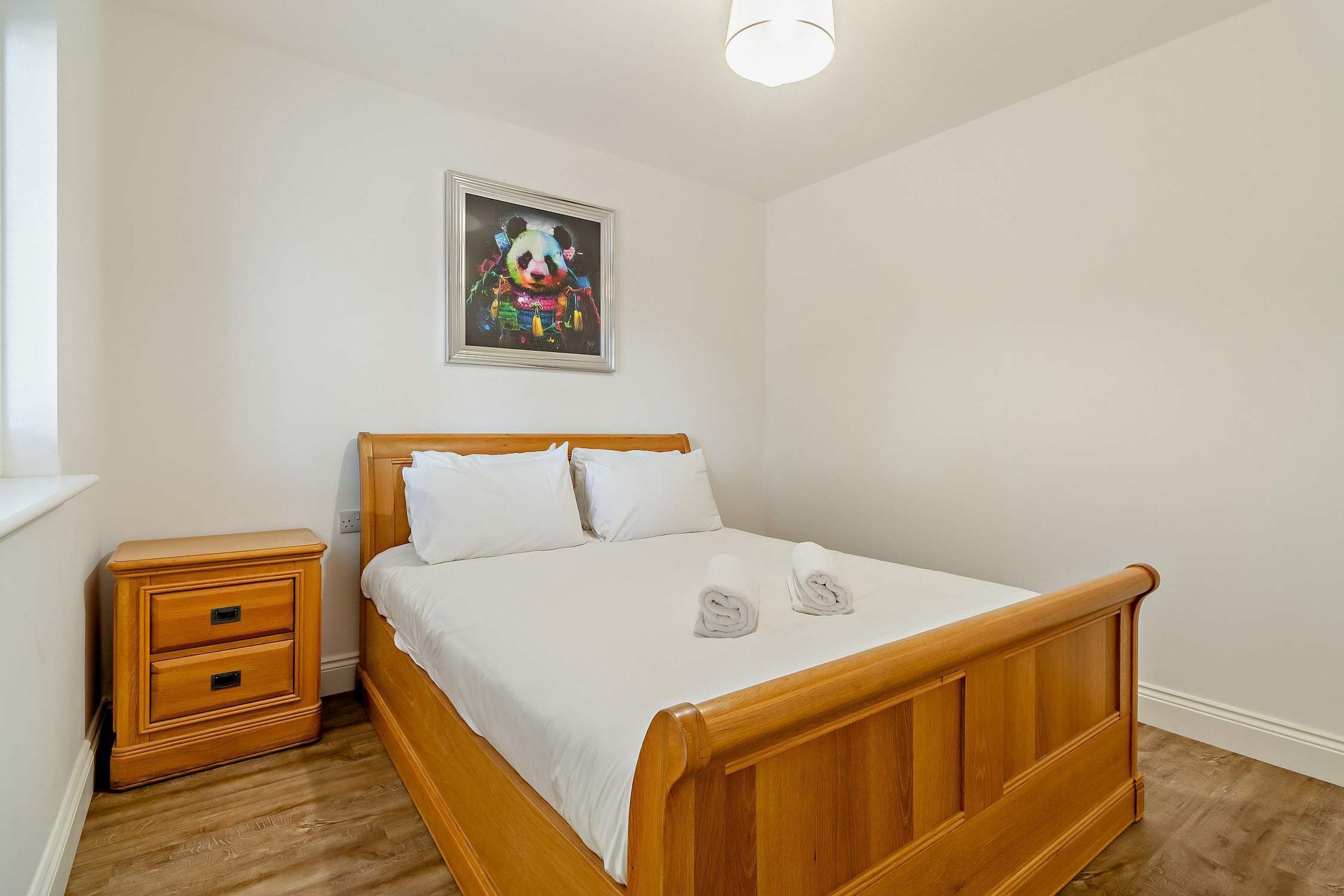 Ein Schlafzimmer mit einem hölzernen Bett, weißer Bettwäsche, einem Nachttisch, einer Schublade und einem an der Wand hängenden Gemälde.