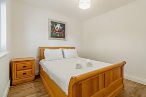 A bedroom with a wooden bed, white bedding, a nightstand, a drawer, and a wall-mounted painting. - Host Stay Robin Lodge (Great Yarmouth)