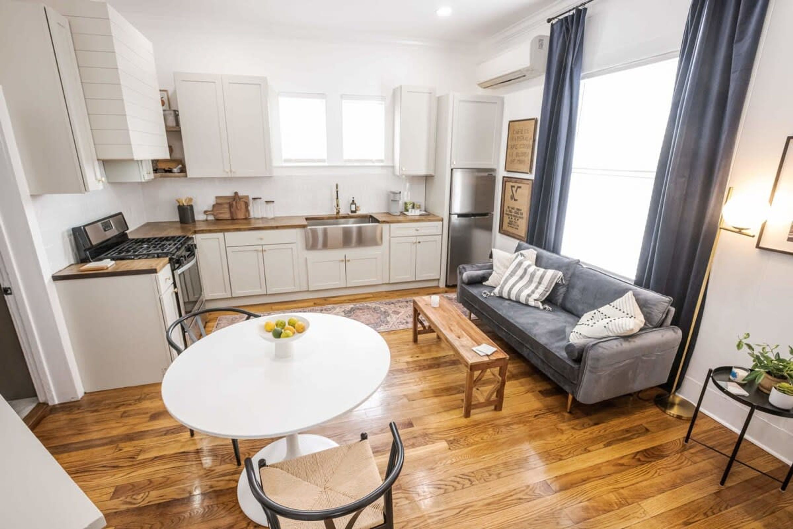 A modern kitchen with white cabinets, a stainless steel refrigerator, and a wooden countertop. There is a gray sofa with a patterned throw pillow and a small table with a bowl of fruit.