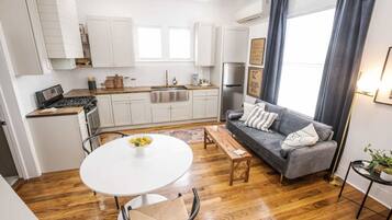 A modern kitchen with white cabinets, a stainless steel refrigerator, and a wooden countertop. There is a gray sofa with a patterned throw pillow and a small table with a bowl of fruit.