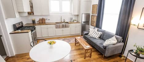 A modern kitchen with white cabinets, a stainless steel refrigerator, and a wooden countertop. There is a gray sofa with a patterned throw pillow and a small table with a bowl of fruit.