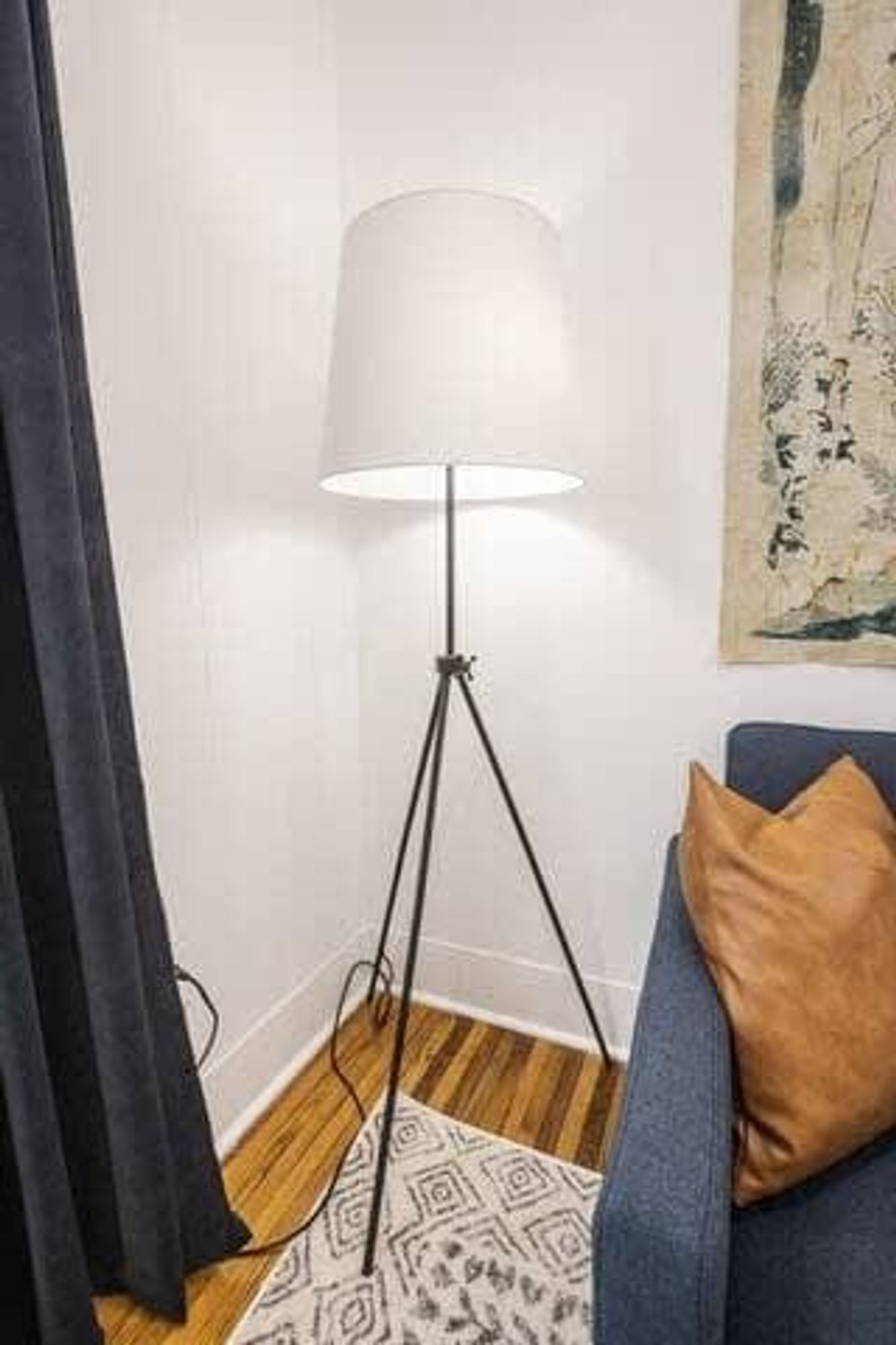 A floor lamp with a white shade, a blue sofa with a brown pillow, and a patterned rug on a wooden floor.