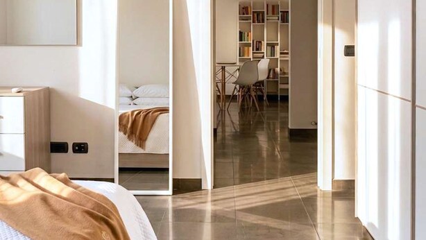 A modern hotel room with a bed, bedside table, and a large mirror. There is a wardrobe, a door leading to a dining area, and a bookshelf in the background. - Casa Bonvicino Citt Studi (Milan)