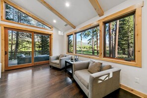 A spacious room with a wooden floor, a sofa, a small table, and large windows offering a view of trees and a deck. - The Alpine Green Lodge (South Lake Tahoe)