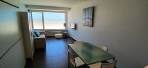 Quarto compacto com mesa de jantar, cadeiras, sofá, radiador e uma grande janela com vista para a praia.