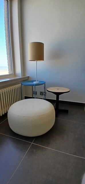 Ein modernes Zimmer mit einem runden, weißen Pouf, einem kleinen blauen Tisch mit Lampe und Blick auf das Meer durch ein Fenster.