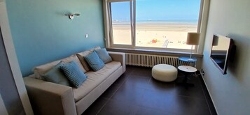 Una habitación con un sofá color beige, una mesita redonda pequeña, un televisor y vistas a la playa.