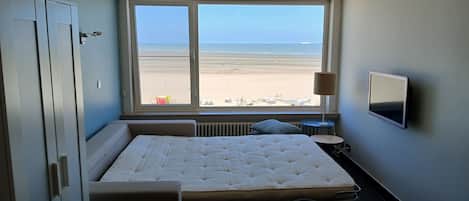 A bedroom with a large window, a bed, a bedside table, a lamp, and a TV.