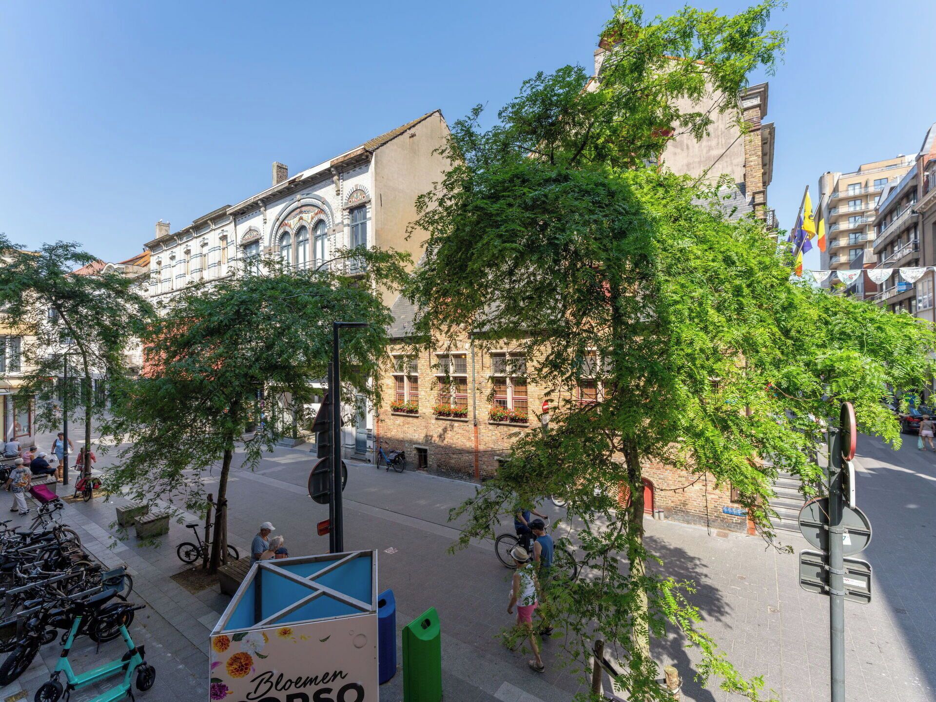 Straatbeeld met historische gebouwen, bomen en een blauw bord waarop 'Bloemen' staat.