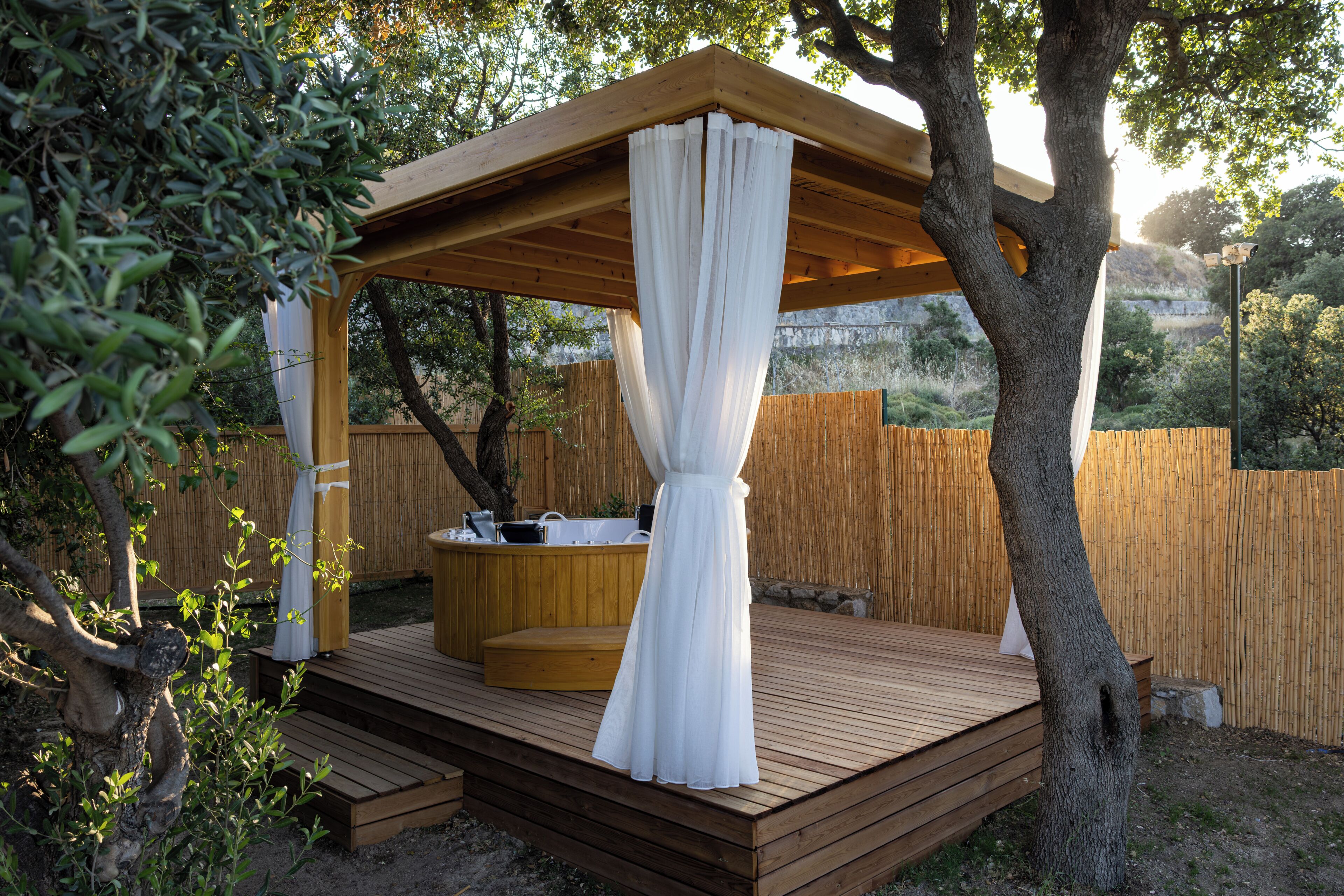 Una terraza de madera con un jacuzzi, una pérgola con cortinas blancas y una valla de madera al fondo.