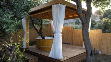 Una terraza de madera con un jacuzzi, una pérgola con cortinas blancas y una valla de madera al fondo.