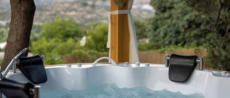 Un jacuzzi con dos sillas negras, rodeado por una terraza de madera y un árbol, con vista a zonas verdes y colinas al fondo.