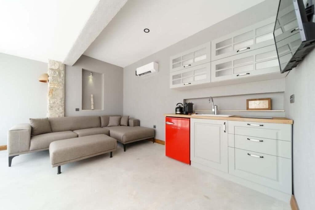 Une cuisine moderne avec des meubles blancs et un appareil électroménager rouge, un canapé d’angle beige et un mur d’accent en pierre.