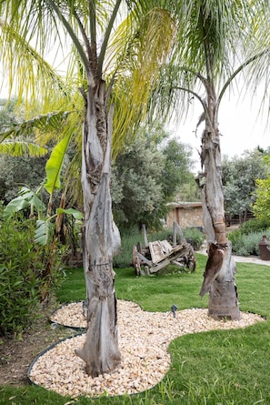 A garden with two palm trees, a stone building, and a wooden cart.