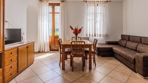 Een kamer met een eettafel, stoelen, een bank, een televisie op een kast en een raam met gordijnen.