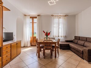 Ein Zimmer mit Esstisch, Stühlen, einer Couch, einem Fernseher auf einem Schrank und einem Fenster mit Vorhängen.