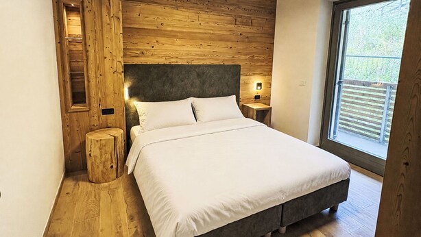 Ein Schlafzimmer mit einem hölzernen Kopfteil, einem Bett mit weißer Bettwäsche, einem Nachttisch mit Lampe und einem Fenster mit Blick ins Grüne.